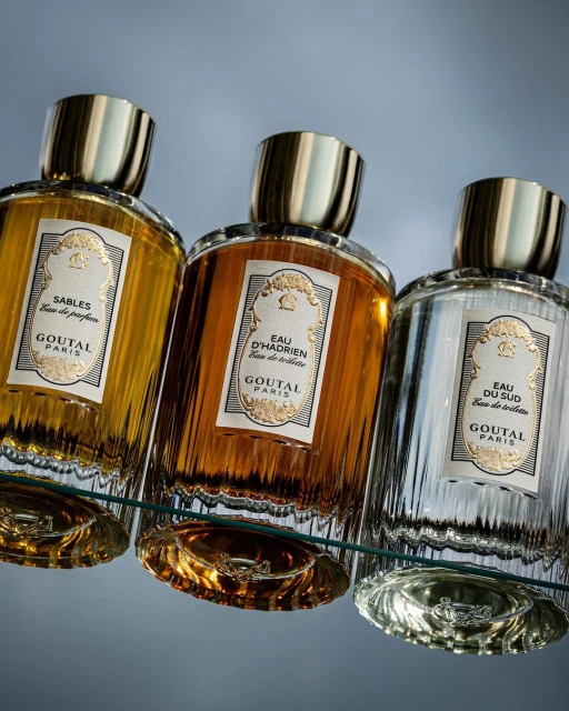 Three Goutal Paris perfume bottles in amber, gold and silver tones are displayed on a clear glass shelf against a gradient gray background.