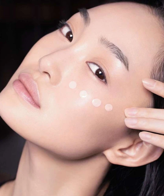 Close-up portrait of a woman with fair skin, wearing beauty product dots on her face, finger to her cheek, and a dark background.