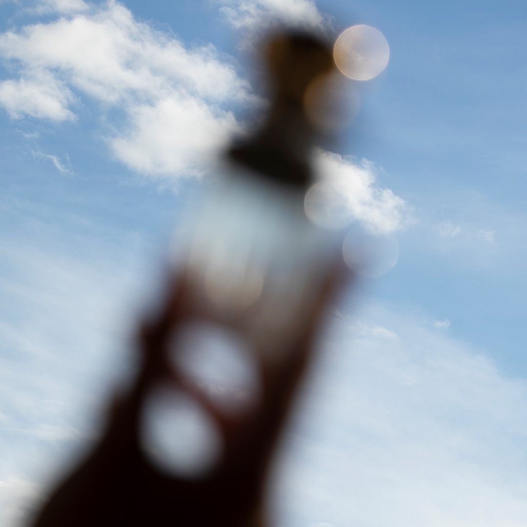 Blurred image of a bottle against a bright blue sky with scattered clouds creates an abstract, dreamlike quality.