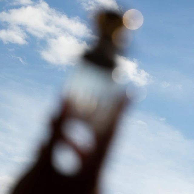 Blurred image of a bottle against a bright blue sky with scattered clouds creates an abstract, dreamlike quality.