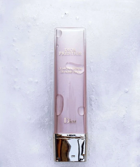 Dior Prestige Le Micro-Serum de Rose Yeux bottle on a textured white surface with water droplets, showcasing beauty product.