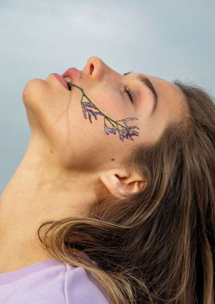 Woman with closed eyes and lavender branch detail across her face in a beauty shot with natural makeup, soft diffused lighting, indoors.