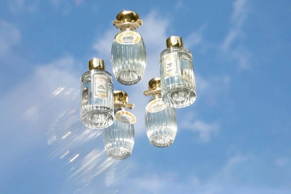 Five perfume bottles with gold caps floating in front of a blue sky with scattered clouds and bright sunlight creating lens flares.