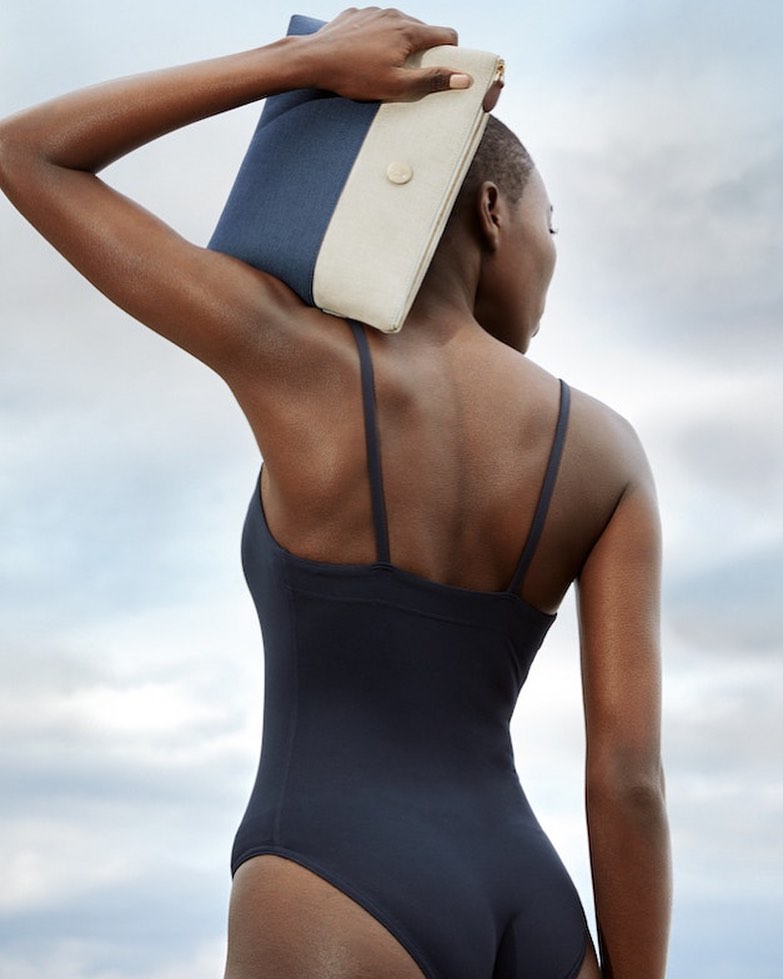 Woman in a black swimsuit stands with her back to the camera, holding a blue and beige clutch purse against a cloudy sky.