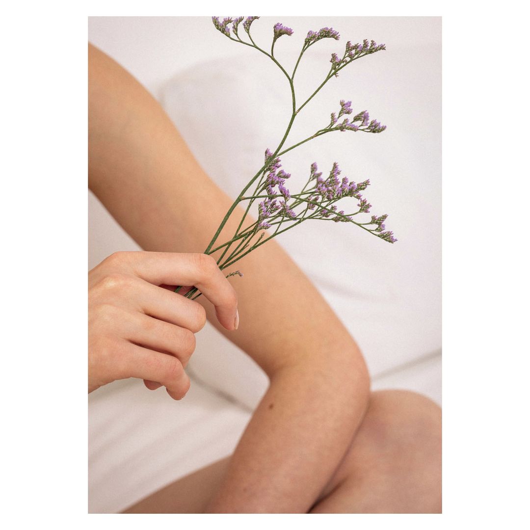 Woman's arm and hand holding sprigs of small purple flowers against a blurred white background, studio lighting. Beauty aesthetic.