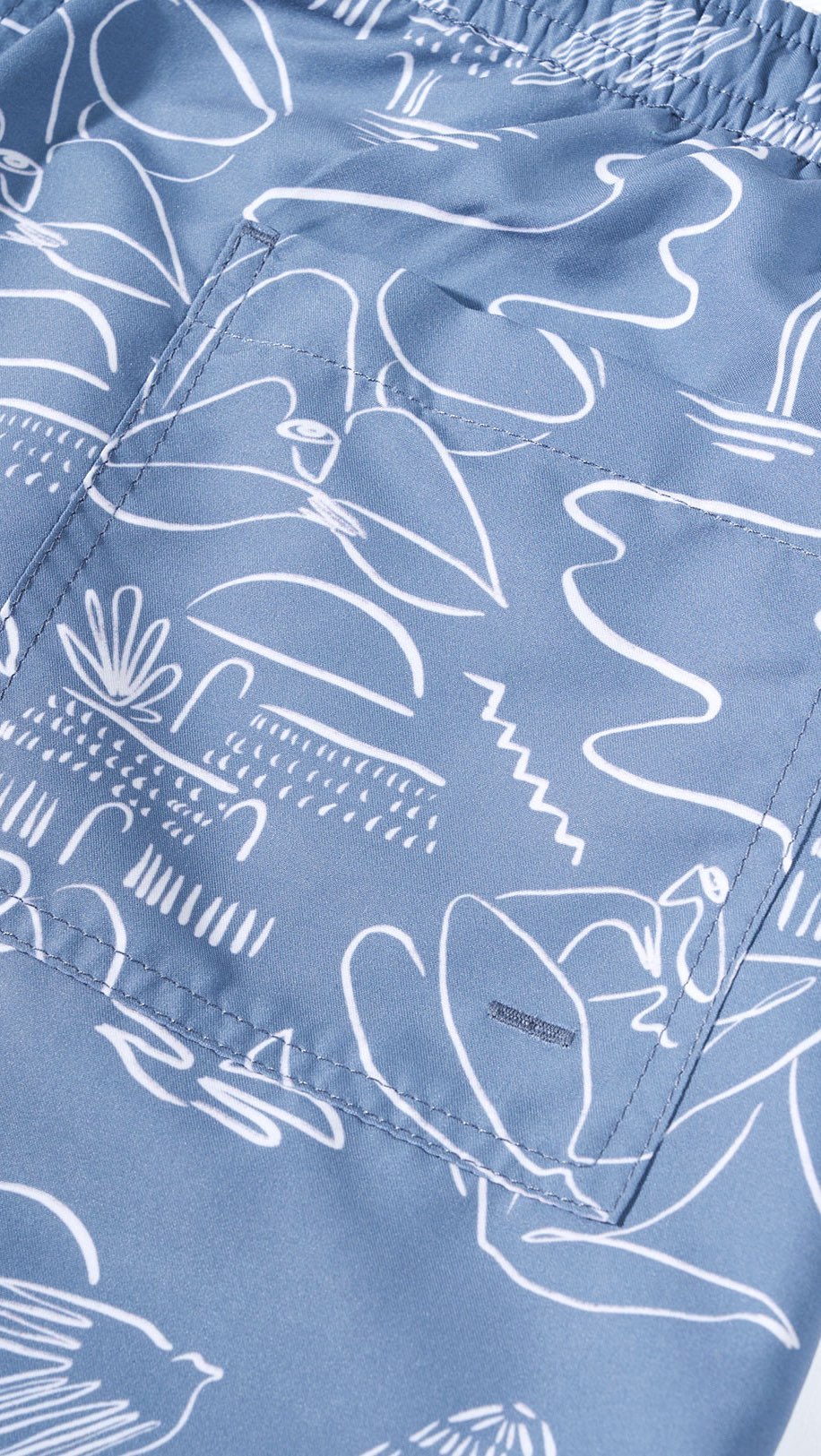 Close-up on blue swim trunks featuring a white line art pattern with tropical island, beach, and ocean motifs in a repeating design.