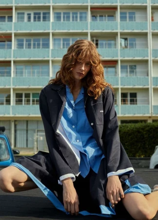 Model with wavy brown hair wearing a blue button-down shirt and dark jacket against a residential building exterior