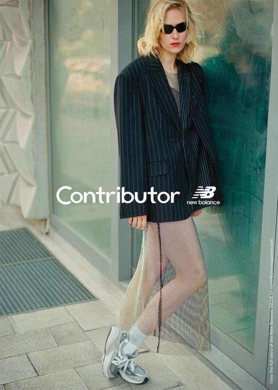 Woman in pinstripe blazer and mesh skirt poses with New Balance sneakers in an urban setting with geometric wall pattern.
