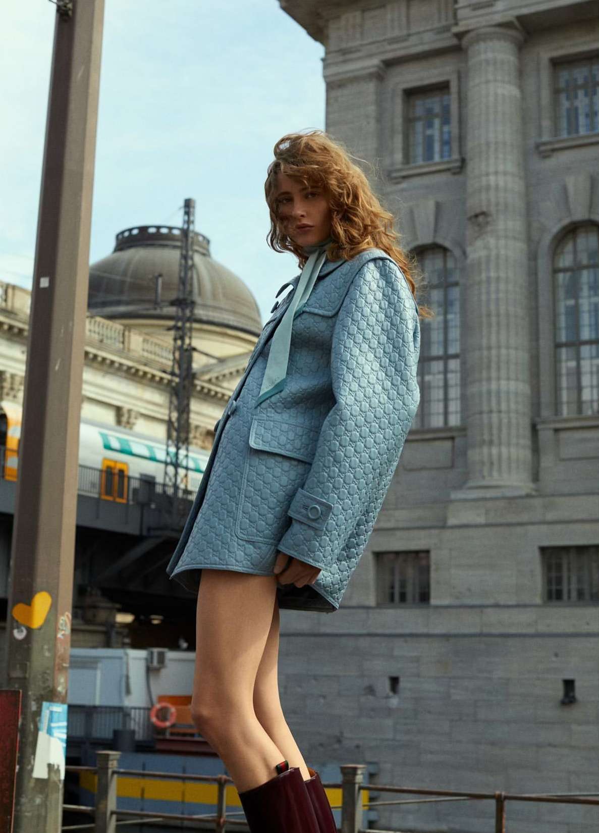 Fashion shot of a fair-skinned adult female with wavy auburn hair wearing a patterned blue jacket and maroon boots, standing outdoors.
