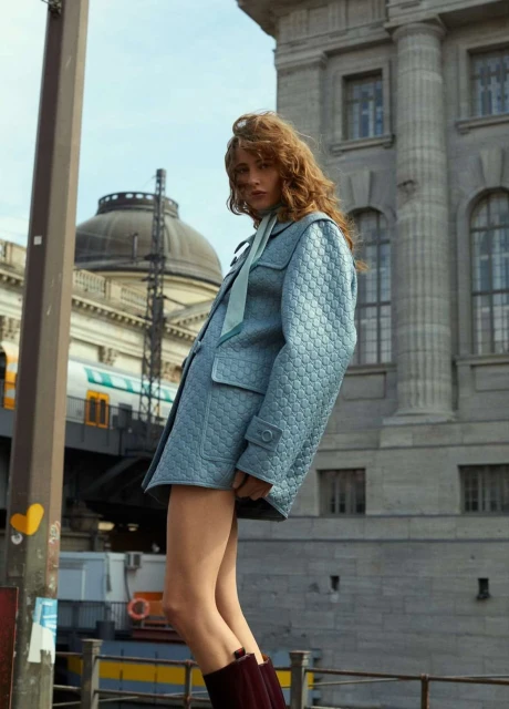 Fashion shot of a fair-skinned adult female with wavy auburn hair wearing a patterned blue jacket and maroon boots, standing outdoors.