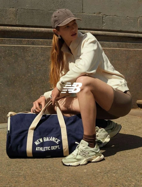 A young woman in activewear poses outdoors with a New Balance duffel bag and sneakers, showcasing athletic fashion and lifestyle.