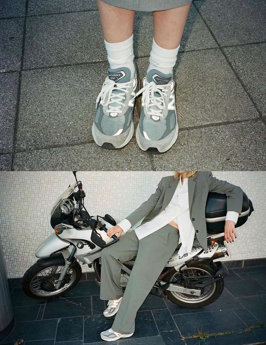 Eye-level shot showcasing gray New Balance sneakers worn with white socks and a gray skirt, alongside a person in a matching suit and motorcycle.