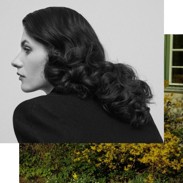 Portrait of a brunette woman with wavy hair in a black top posing in front of a window and garden with yellow flowers.