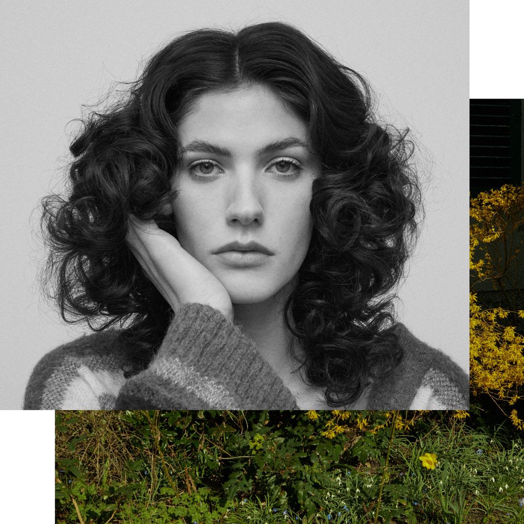 Monochrome portrait of an adult female with curly dark hair, resting her face on her hand, wearing a patterned sweater, in an outdoor setting.