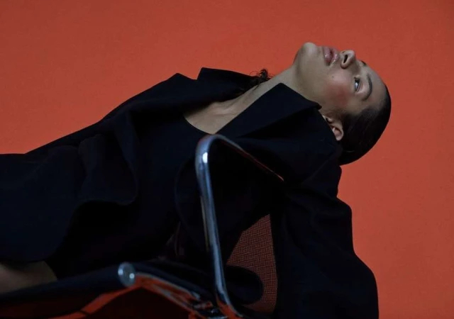 Woman in black blazer leaning back in a chrome chair against a vibrant solid orange seamless studio backdrop