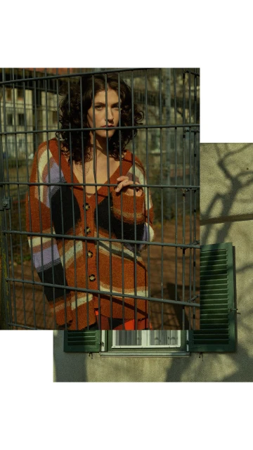 Adult female with curly brown hair, wearing a patterned cardigan, standing behind metal bars, with buildings and trees in the background.