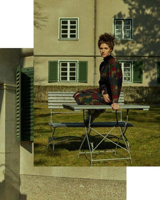 A woman sits on a metal bench in a garden setting with a retro-inspired floral dress, curly hair, and a neutral expression outdoors.