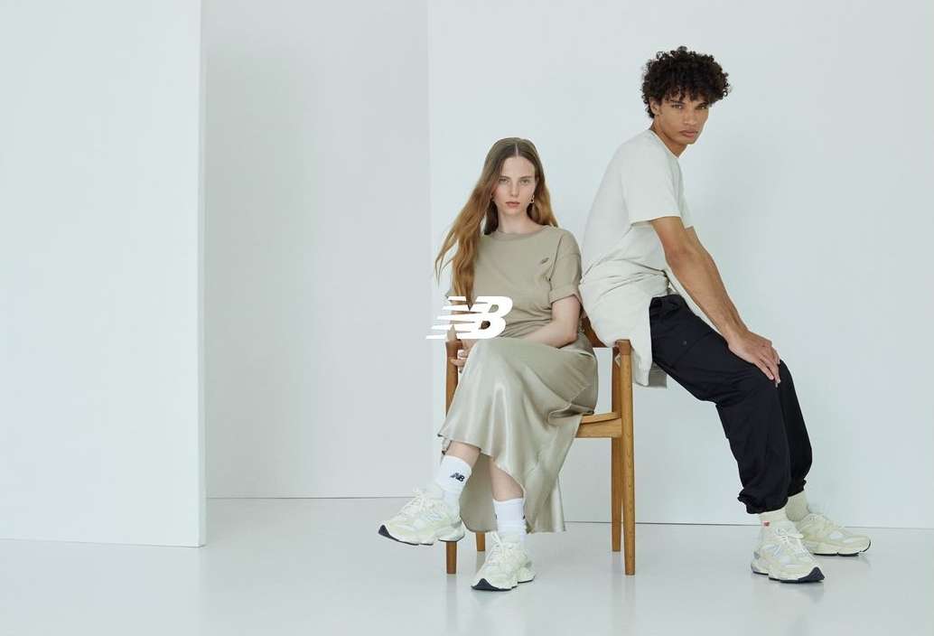 Two models wearing beige shirts and sneakers sit indoors on a wooden chair in a minimalist photo studio with a white background.
