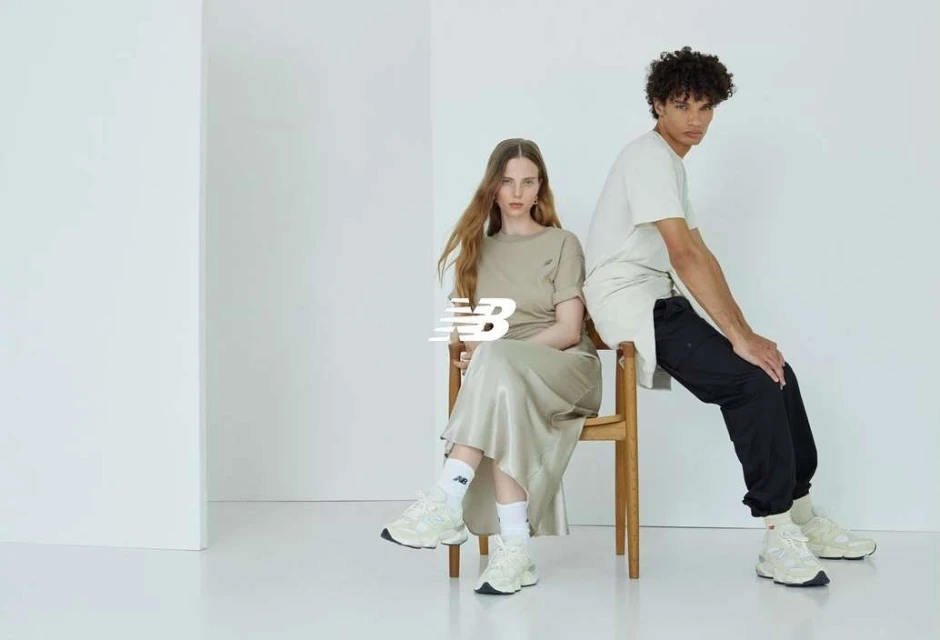 Two models wearing beige shirts and sneakers sit indoors on a wooden chair in a minimalist photo studio with a white background.