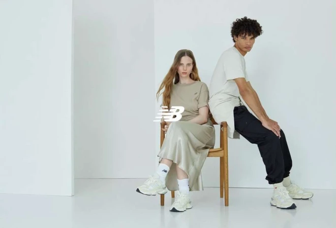 Two models wearing beige shirts and sneakers sit indoors on a wooden chair in a minimalist photo studio with a white background.