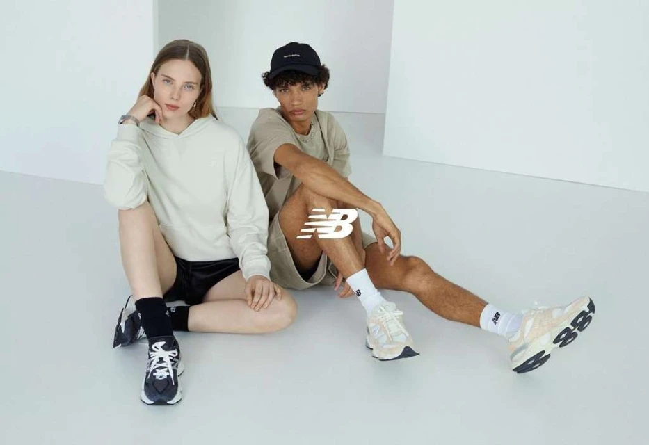 Two fashion models pose in a minimalist studio setting showcasing athletic sneakers and streetwear clothing, displaying the 'New Balance' logo.