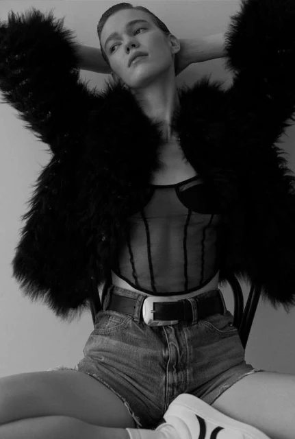 Black-and-white portrait of a female model wearing a faux fur jacket, denim shorts, a sheer corset top, and sneakers, sitting on a chair.