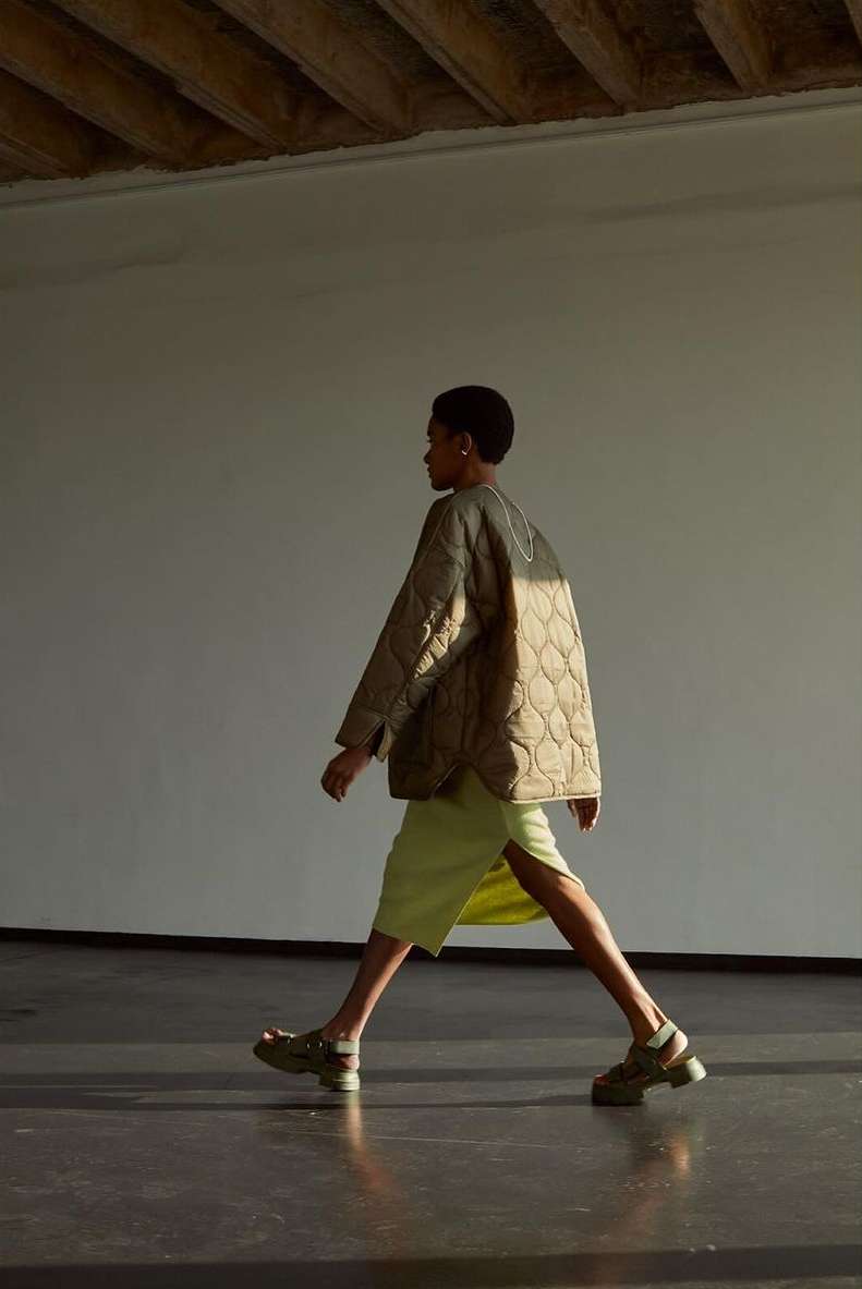 Side view of a Black female model walking and posing indoors. She wears a tan quilted jacket, light green dress, and chunky sandals.