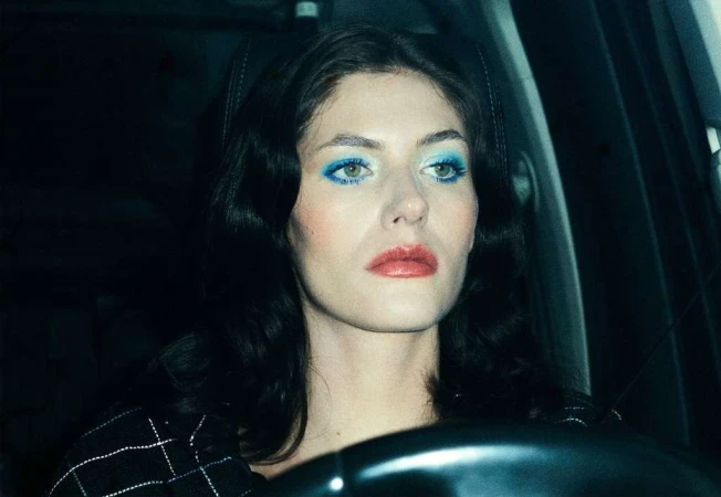 Close-up portrait of a dark-haired woman with blue eyeshadow driving at night, steering wheel visible, fashion, and automotive photography.