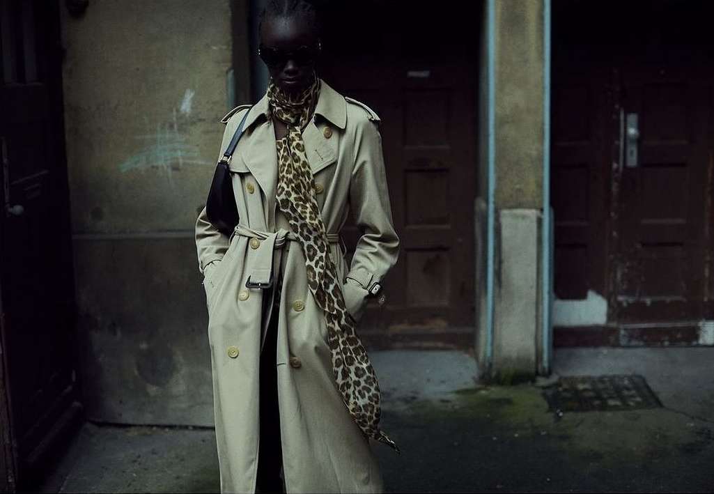 Fashionable woman in a trench coat and leopard-print scarf stands in an urban doorway with neutral tones and a shadowed ambiance.