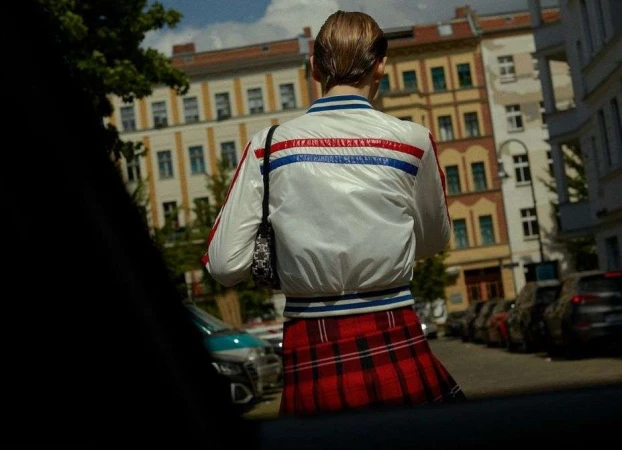 Model seen from behind wearing a white bomber jacket and red plaid skirt standing on a sunny city street.