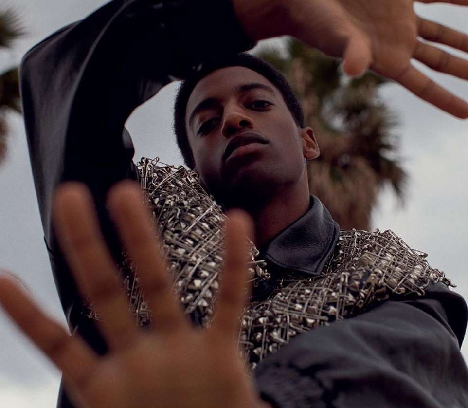 A stylish African-American man in a black leather jacket with pin embellishments reaches toward the camera, posing outdoors with palm trees.