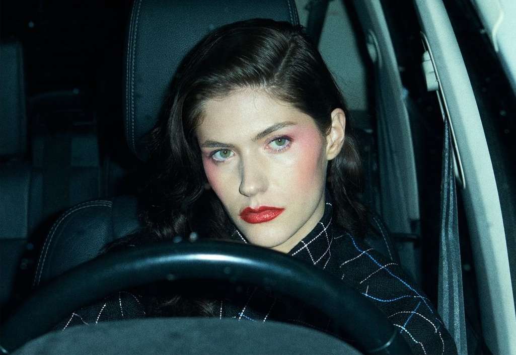 Close-up of a woman with vivid makeup sitting in a car, focusing on her face and expression, with the steering wheel visible.