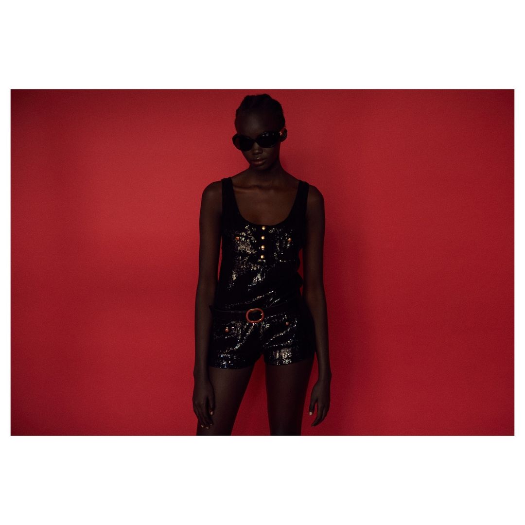 A Black woman poses in a glossy black romper with gold button accents against a solid red backdrop, highlighting fashion and portrait photography.