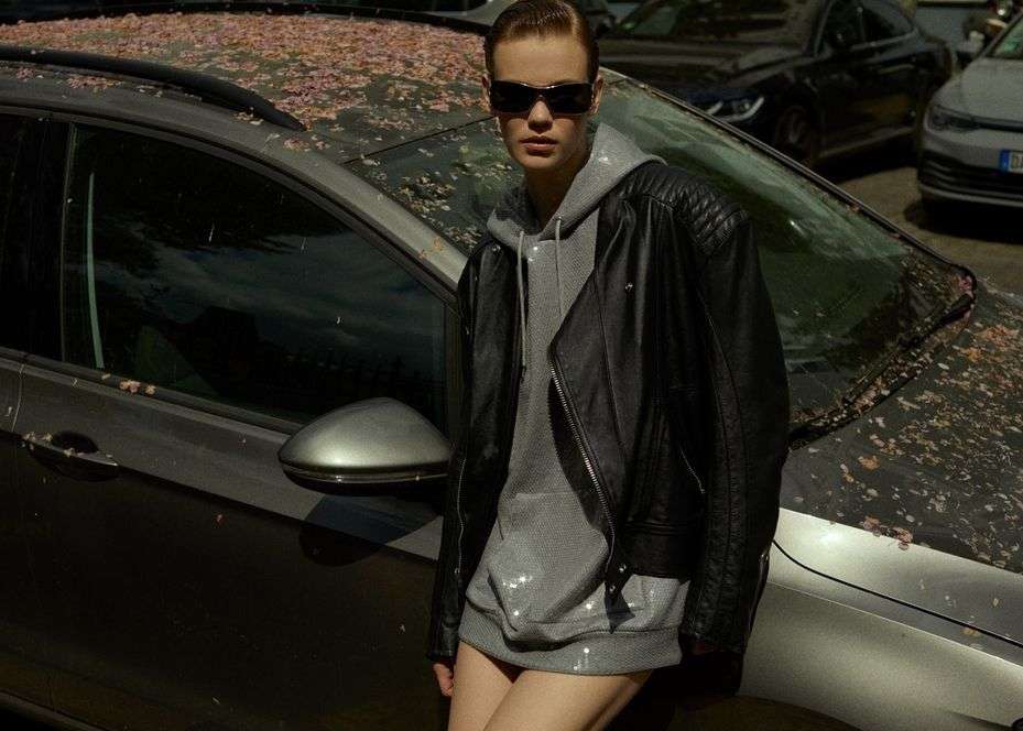 Woman in sunglasses, leather jacket, and hoodie poses next to a grey car covered in pink petals in an urban environment.