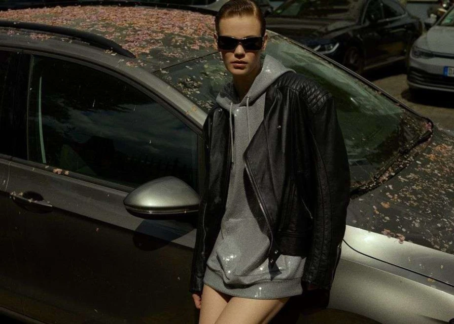 Woman in sunglasses, leather jacket, and hoodie poses next to a grey car covered in pink petals in an urban environment.