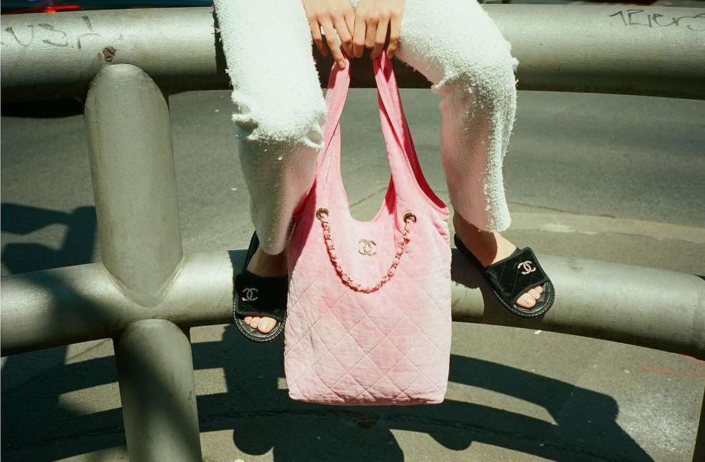 Fashion still life features a pink Chanel velvet tote bag on person's lap wearing fuzzy pants and Chanel logo slide sandals on a metal structure.