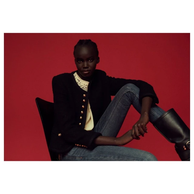A woman with dark skin sits in a black chair against a red backdrop, wearing a black jacket, light jeans, and black boots in a studio setting.