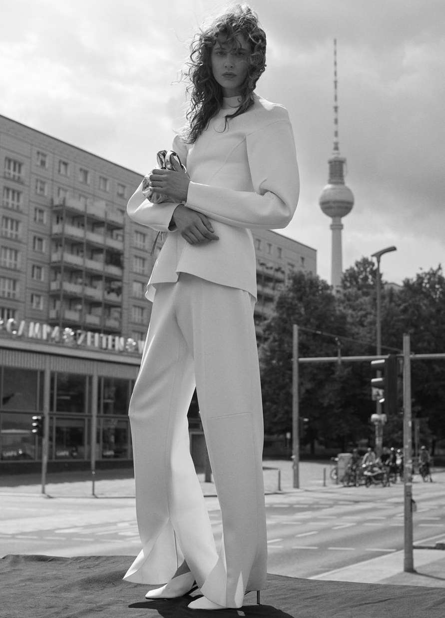 A woman in a white suit poses on a city street in front of buildings and a radio tower under a cloudy sky in a black and white fashion shot.