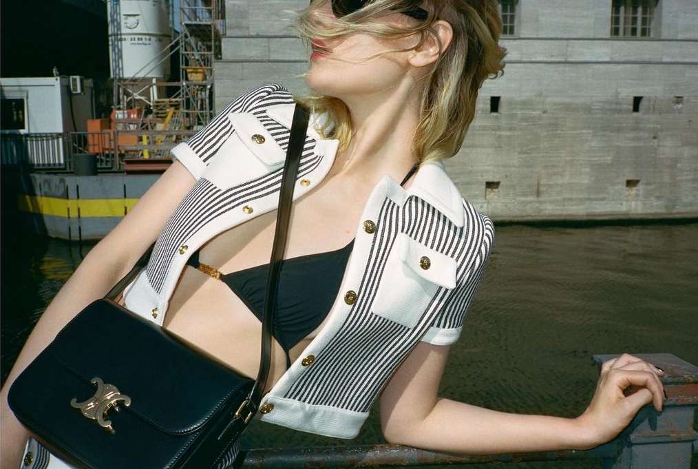 A woman in a striped jacket and black bikini top poses outdoors with a Celine purse against a waterway and building backdrop.