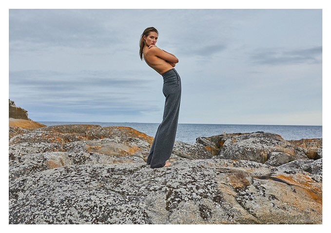 Fashion shot of a woman posing on rocky terrain, wearing gray pants against an ocean and sky backdrop on a cloudy day.