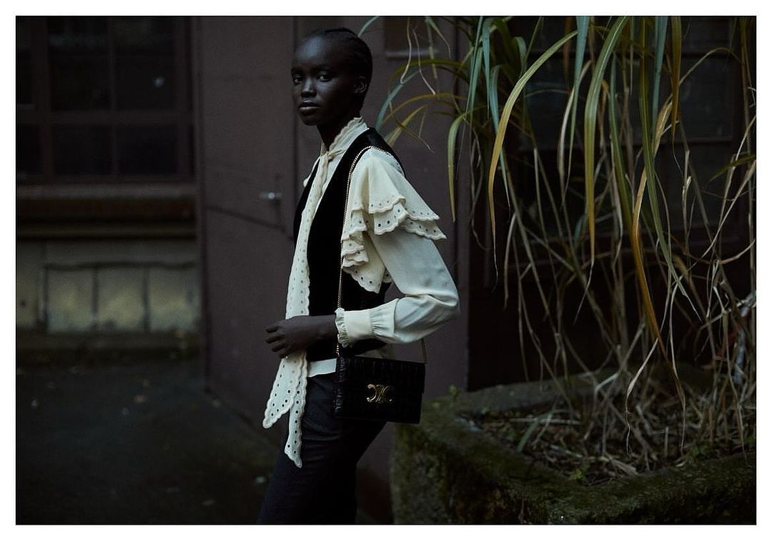 Adult female model wearing black vest, white blouse with ruffles, and black purse with logo in outdoor fashion editorial setting.