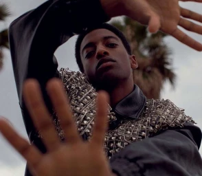 Black man wearing a black leather jacket covered in safety pins reaching toward the camera with out-of-focus palm