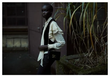 Black woman wearing a ruffled cream blouse and dark vest holding a black shoulder bag outdoors by foliage.