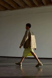 Model in profile wearing a quilted olive jacket and light green skirt walking across a sunlit room by Magnus Magnusson