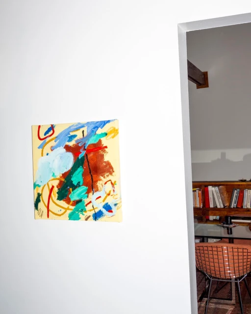 Abstract painting on a white wall in an interior setting, with a view through a doorway to a chair and bookshelf.
