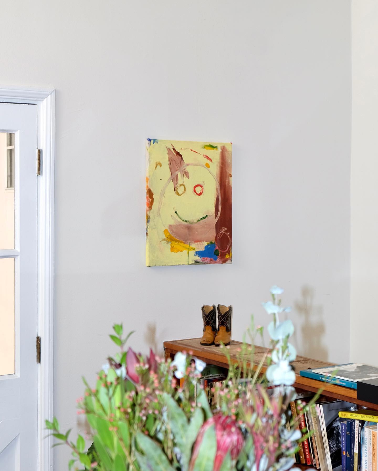 Abstract art featuring a smiley face hangs above cowboy boots on a shelf, with flowers visible in the foreground in a home interior.