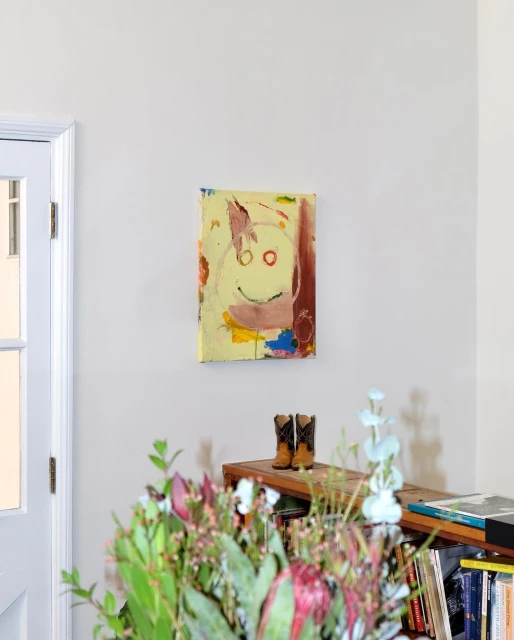 Abstract art featuring a smiley face hangs above cowboy boots on a shelf, with flowers visible in the foreground in a home interior.