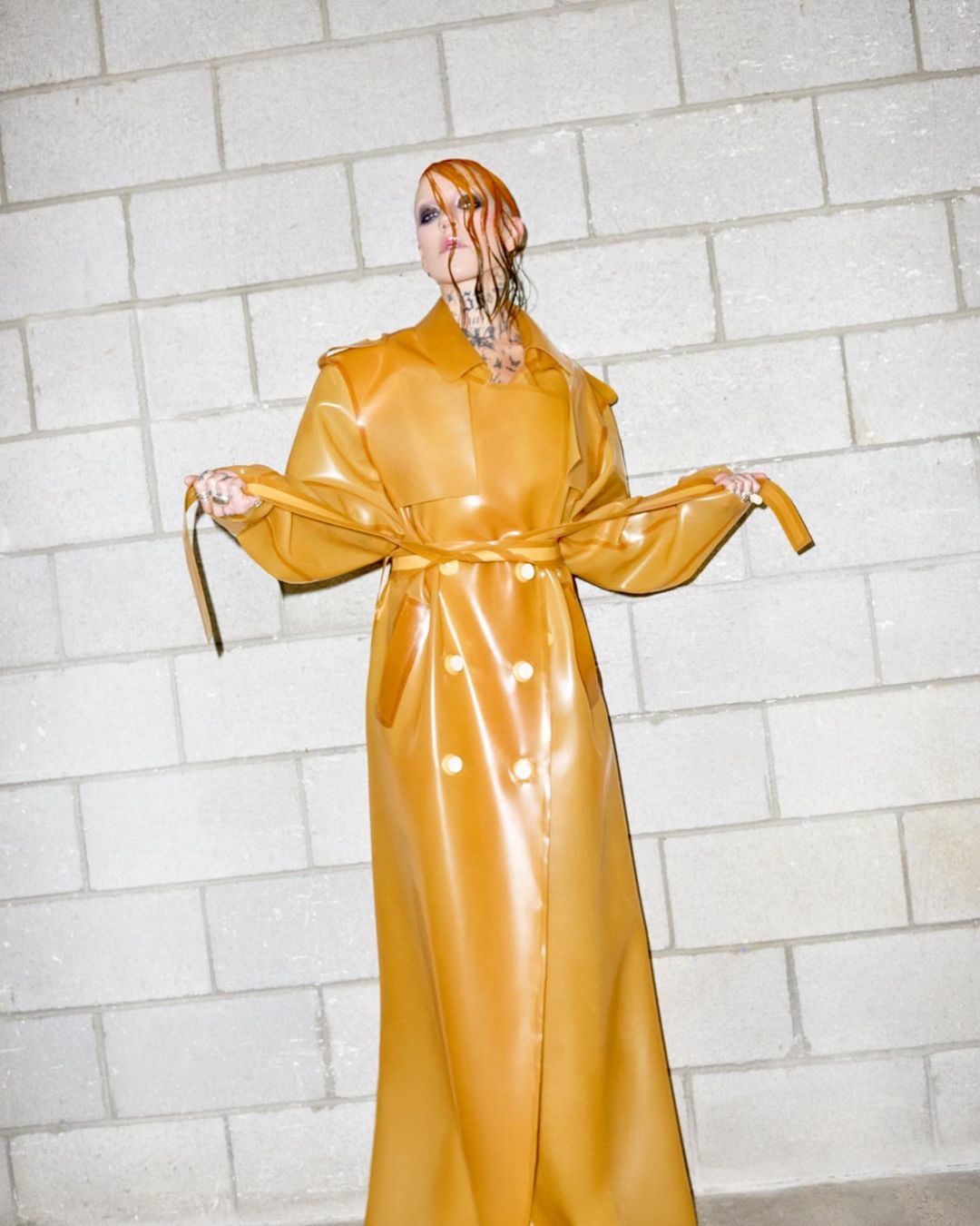 Adult female model with wet auburn hair poses in a shiny golden double-breasted trench coat in front of a light-gray brick wall.