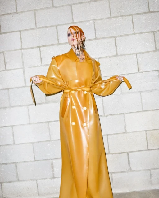 Adult female model with wet auburn hair poses in a shiny golden double-breasted trench coat in front of a light-gray brick wall.