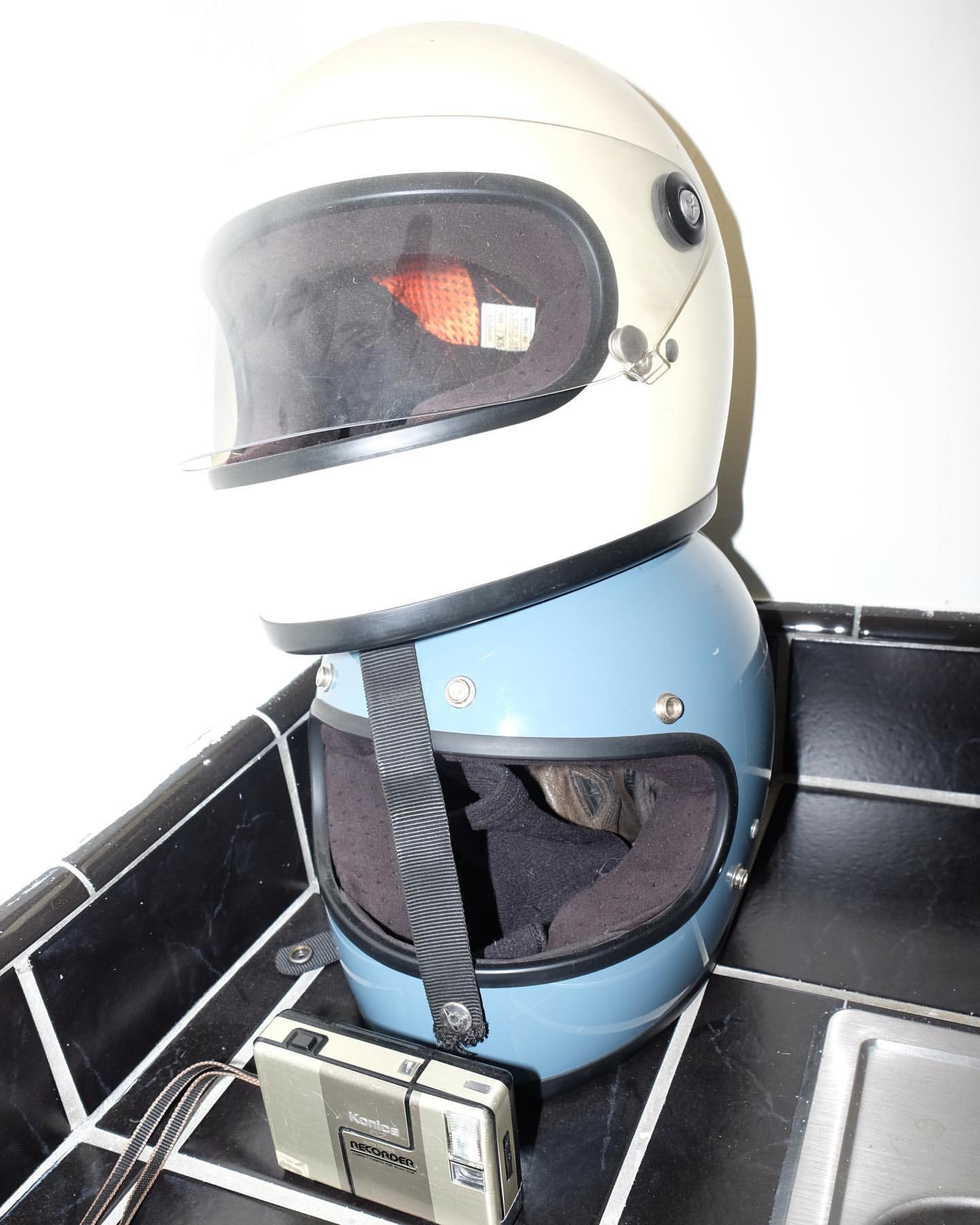 Stacked motorcycle helmets in white and blue, with a vintage camera resting on black-and-white tiled surface.